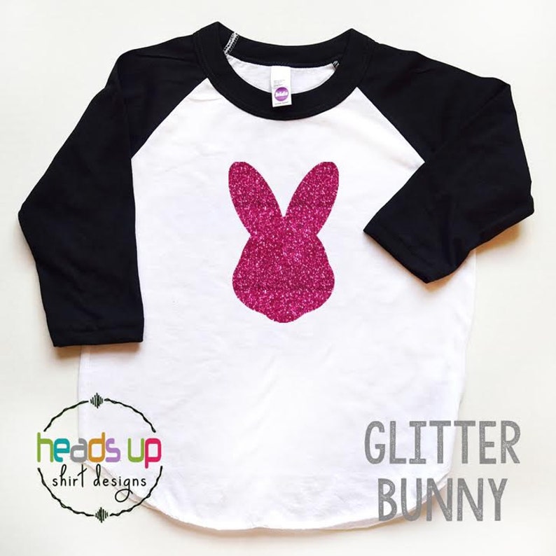 May include: A white and black baseball tee shirt with a pink glitter bunny silhouette on the front. The shirt has the text "GLITTER BUNNY" printed in gray below the bunny. The text "heads up shirt designs" is printed in a circle on the bottom left of the shirt.