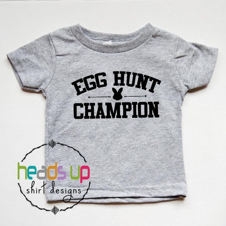 Puede incluir: Camiseta gris de manga corta con texto negro que dice "Egg Hunt Champion" con un gr&aacute;fico de conejo.