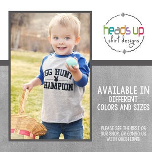 Peut inclure: Un jeune enfant portant un t-shirt de baseball gris et bleu avec le texte "Egg Hunt Champion" et un graphique de lapin. L'enfant tient un &oelig;uf de P&acirc;ques bleu et un panier en osier.