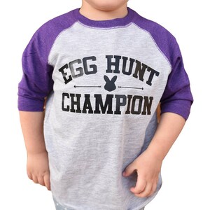 Peut inclure: Un t-shirt de baseball gris et violet avec le texte "Egg Hunt Champion" et un graphique de lapin.