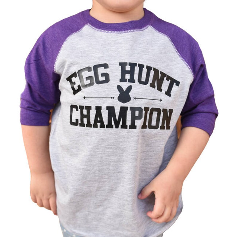 Peut inclure: Un t-shirt de baseball gris et violet avec le texte "Egg Hunt Champion" et un graphique de lapin.