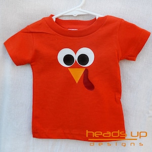 May include: Orange short-sleeved t-shirt with a turkey face design. The turkey has large white eyes, an orange beak, and a red wattle.