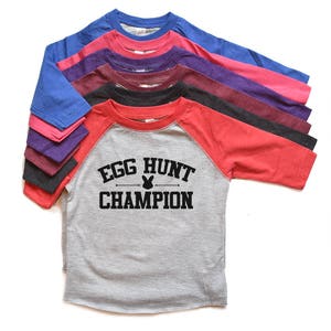 Peut inclure: Une pile de huit t-shirts de baseball de diff&eacute;rentes couleurs, dont rose, violet, bleu, rouge et gris. Le t-shirt gris porte le texte "Egg Hunt Champion" avec un graphique de lapin.