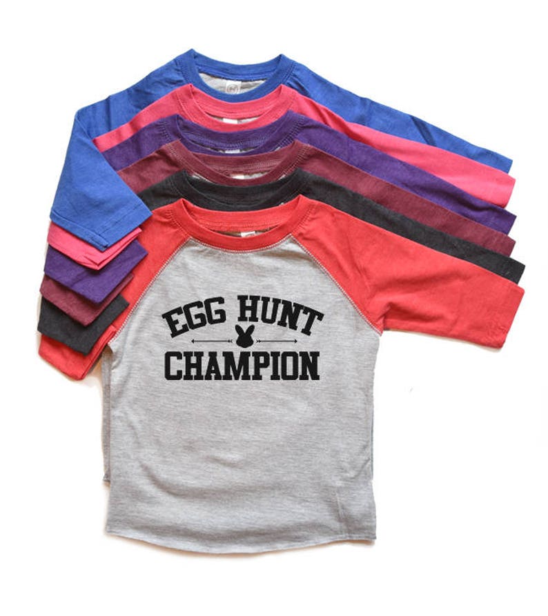 Peut inclure: Une pile de huit t-shirts de baseball de diff&eacute;rentes couleurs, dont rose, violet, bleu, rouge et gris. Le t-shirt gris porte le texte "Egg Hunt Champion" avec un graphique de lapin.