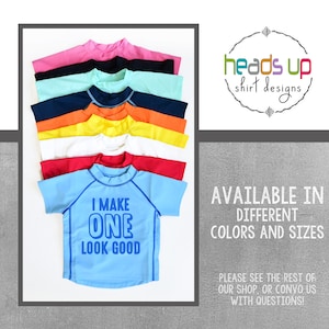 Peut inclure: Une pile de dix chemises à manches courtes de différentes couleurs, dont rose, bleu, orange, jaune et blanc. La chemise du bas est bleu clair avec le texte "I MAKE ONE LOOK GOOD" en bleu.