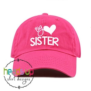 Puede incluir: Gorra de béisbol rosa con texto blanco que dice "Big Sister" con un símbolo de corazón.