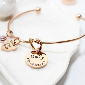 May include: Rose gold bangle bracelet with a knot detail and two charms. One charm is a heart with the text "Mother of the Bride" and the other is a round charm with the text "S♥J 02.14.2020".