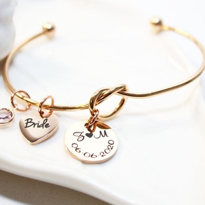 May include: Gold bangle bracelet with a knot design and three charms: a heart charm with the word "Bride" engraved on it, a round charm with the initials "J & M" and the date "06-06-2020" engraved on it, and a small clear crystal charm.