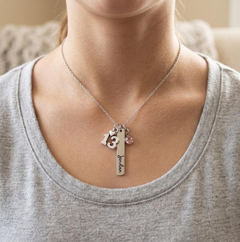 May include: A silver-toned necklace featuring a vertical rectangular bar pendant engraved with the name Jordan in black script, a charm shaped like the number 13 with a small heart, and a single round pink crystal charm on a delicate chain.
