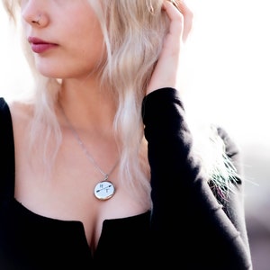Puede incluir: Un collar plateado con un colgante redondo con un dise&ntilde;o en blanco y negro de flechas y l&iacute;neas. El colgante cuelga de una cadena delicada. La persona lleva una camiseta negra.