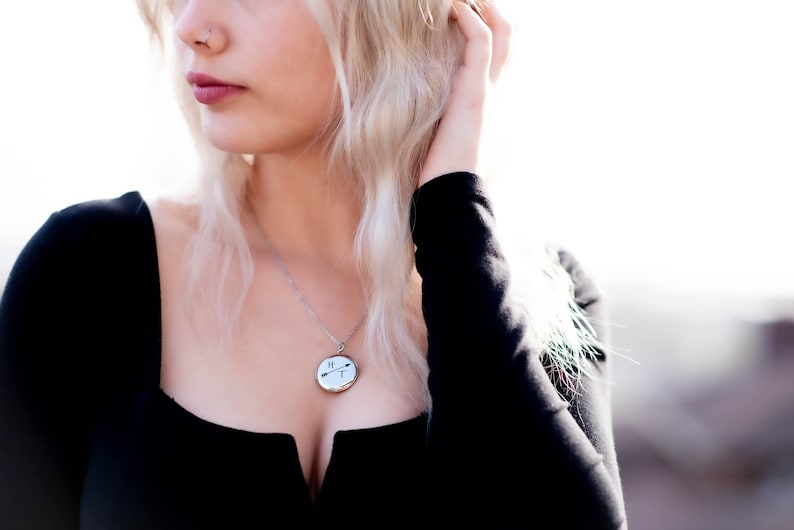 Puede incluir: Un collar plateado con un colgante redondo con un dise&ntilde;o en blanco y negro de flechas y l&iacute;neas. El colgante cuelga de una cadena delicada. La persona lleva una camiseta negra.