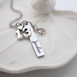 Puede incluir: Collar de plata con un dije que dice "13" y un dije de barra con el nombre "Jordan" grabado. El collar tambi&eacute;n tiene un peque&ntilde;o dije de coraz&oacute;n y un cristal rosa.
