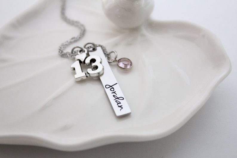 Puede incluir: Collar de plata con un dije que dice "13" y un dije de barra con el nombre "Jordan" grabado. El collar tambi&eacute;n tiene un peque&ntilde;o dije de coraz&oacute;n y un cristal rosa.