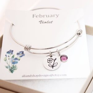 May include: A silver expandable bangle bracelet with a charm featuring a violet flower and a small purple gemstone. The bracelet is displayed on a white card with the text "February Violet" and "aliandshaydesigns@etsy.com".
