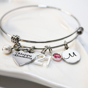 May include: Silver charm bracelet with a heart charm that says "Happy Birthday", a charm with the number "21", a pink crystal charm, and a charm with the letter "M".