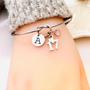 May include: A silver knot bangle bracelet with a pink crystal charm, a charm with the letter "A" and a charm with the number "17".