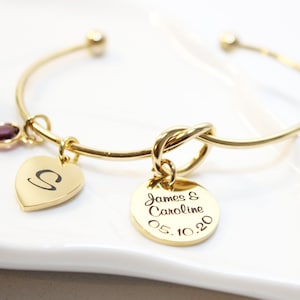 May include: Gold bangle bracelet with a knot charm, a heart charm with the letter "S" engraved on it, and a round charm with the text "James & Caroline 05.10.20" engraved on it. A purple gemstone is attached to the bangle.