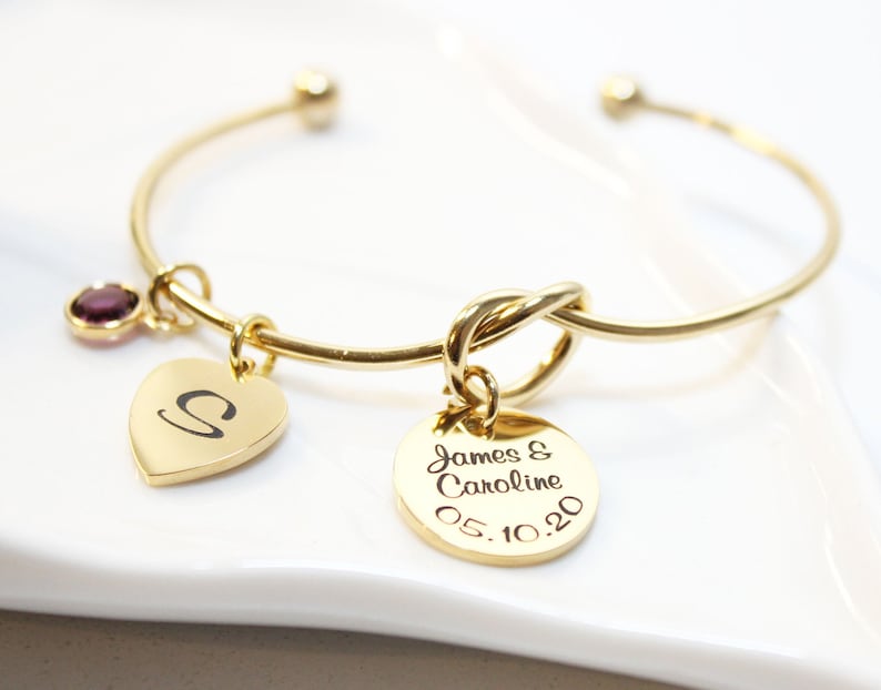 May include: Gold bangle bracelet with a knot charm, a heart charm with the letter "S" engraved on it, and a round charm with the text "James & Caroline 05.10.20" engraved on it. A purple gemstone is attached to the bangle.