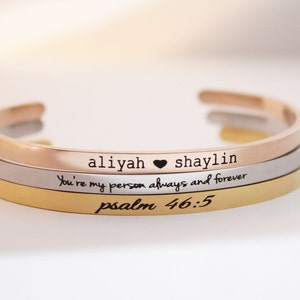 May include: Three open cuff bracelets in rose gold, silver, and gold. The rose gold bracelet has the names "aliyah" and "shaylin" with a heart symbol. The silver bracelet reads "You're my person always and forever". The gold bracelet reads "psalm 46:5".