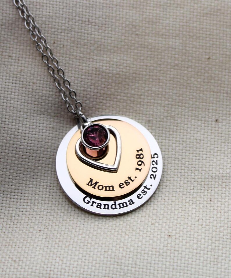 K&ouml;nnte beinhalten: Eine silberne Kettenkette mit einem mehrschichtigen Anh&auml;nger. Der Anh&auml;nger hat einen Herz-Ausschnitt mit einem violetten Edelstein, und die &auml;u&szlig;ere Schicht ist mit "Mom est. 1981 Grandma est. 2025" graviert.