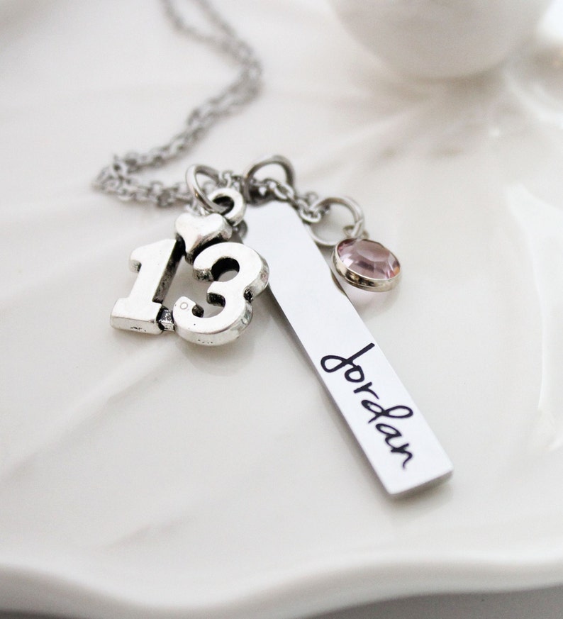 Puede incluir: Collar de plata con un dije que dice "13" y un dije con el nombre "Jordan" grabado. El collar tambi&eacute;n tiene un peque&ntilde;o dije de cristal rosa.