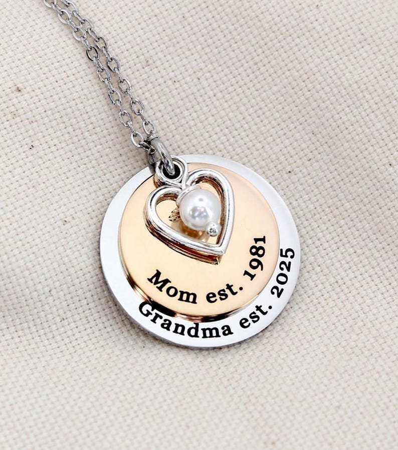 K&ouml;nnte beinhalten: Silberne Halskette mit einem mehrschichtigen Anh&auml;nger, der einen Herz-Anh&auml;nger mit einer Perle und den Text "Mom est. 1981 Grandma est. 2025" tr&auml;gt.