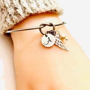 Puede incluir: Pulsera de brazalete plateada con dise&ntilde;o de nudo. La pulsera presenta un dije circular con la letra "A", un dije de ala y una peque&ntilde;a perla. La pulsera se usa en una mu&ntilde;eca, con una manga de su&eacute;ter beige en el fondo.