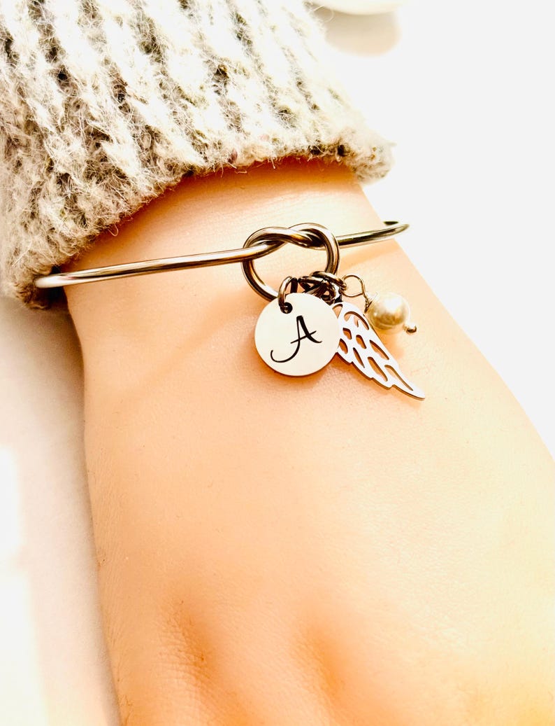 Puede incluir: Pulsera de brazalete plateada con dise&ntilde;o de nudo. La pulsera presenta un dije circular con la letra "A", un dije de ala y una peque&ntilde;a perla. La pulsera se usa en una mu&ntilde;eca, con una manga de su&eacute;ter beige en el fondo.