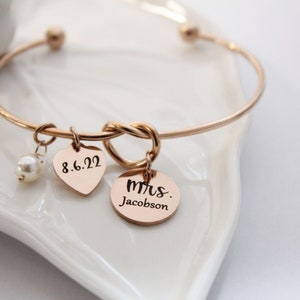 May include: A gold bangle bracelet with a knot design and two charms. One charm is a heart with the date 8.6.22 engraved on it, and the other charm is a round disc with the text "mrs. Jacobson" engraved on it.