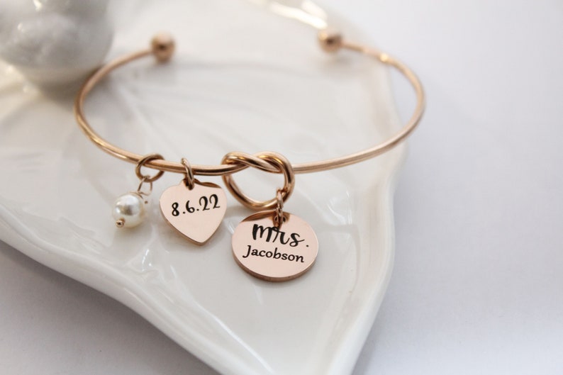 May include: A gold bangle bracelet with a knot design and two charms. One charm is a heart with the date 8.6.22 engraved on it, and the other charm is a round disc with the text "mrs. Jacobson" engraved on it.
