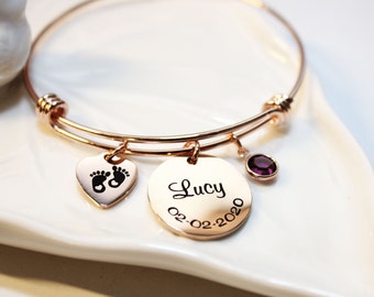 May include: Rose gold bangle bracelet with a heart charm featuring a footprint design, a round charm with the name "Lucy" and the date "02-02-2020" engraved, and a purple gemstone charm.