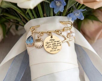 May include: A gold-toned safety pin brooch attached to a white ribbon-wrapped bouquet, featuring a round engraved charm that reads: Lilly, You'll always be our little girl. Love, Mom & Dad 06.20.26, alongside a blue teardrop, seashell, starfish, and pearl.