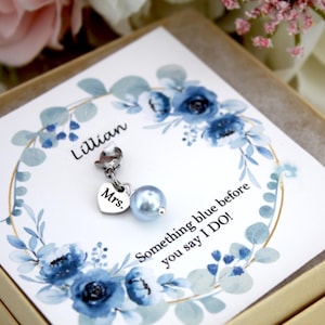 May include: A blue pearl charm with a silver heart pendant that says "Mrs." hangs from a silver clasp. The charm is on a card with a blue floral wreath and the text "Something blue before you say I DO!"