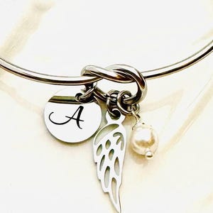 Puede incluir: Una pulsera de brazalete plateada con un dise&ntilde;o de nudo. Cuenta con un dije redondo con la letra "A", un dije de ala calada y una perla falsa colgante. La pulsera es una pieza de joyer&iacute;a delicada.