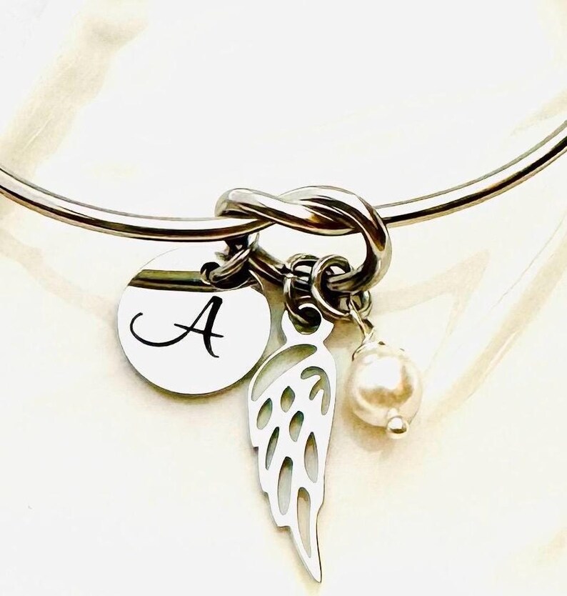 Puede incluir: Una pulsera de brazalete plateada con un dise&ntilde;o de nudo. Cuenta con un dije redondo con la letra "A", un dije de ala calada y una perla falsa colgante. La pulsera es una pieza de joyer&iacute;a delicada.