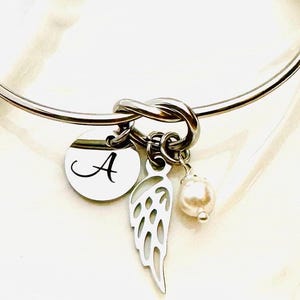 Puede incluir: Una pulsera de brazalete plateada con un dise&ntilde;o de nudo. Cuenta con un dije redondo con la letra "A", un dije en forma de ala y una peque&ntilde;a perla. La pulsera es una joya.