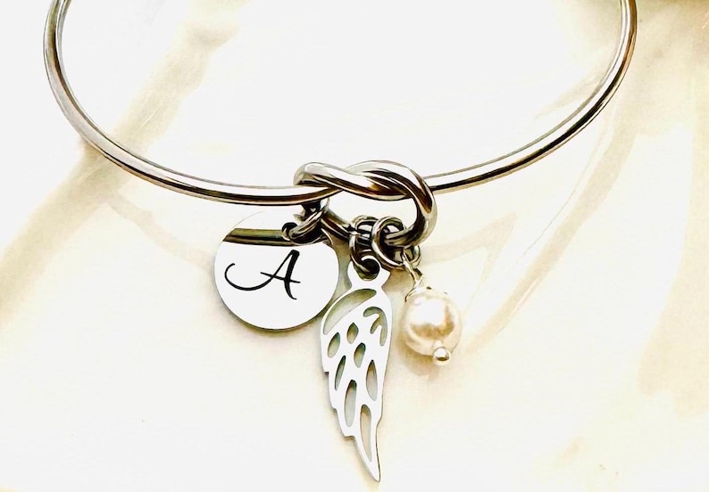 Puede incluir: Una pulsera de brazalete plateada con un dise&ntilde;o de nudo. Cuenta con un dije redondo con la letra "A", un dije en forma de ala y una peque&ntilde;a perla. La pulsera es una joya.