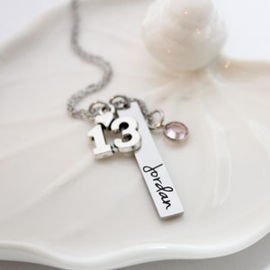 Puede incluir: Collar de plata con un dije que dice "13" y un dije con el nombre "Jordan" grabado. El collar tambi&eacute;n tiene un peque&ntilde;o dije de cristal rosa.