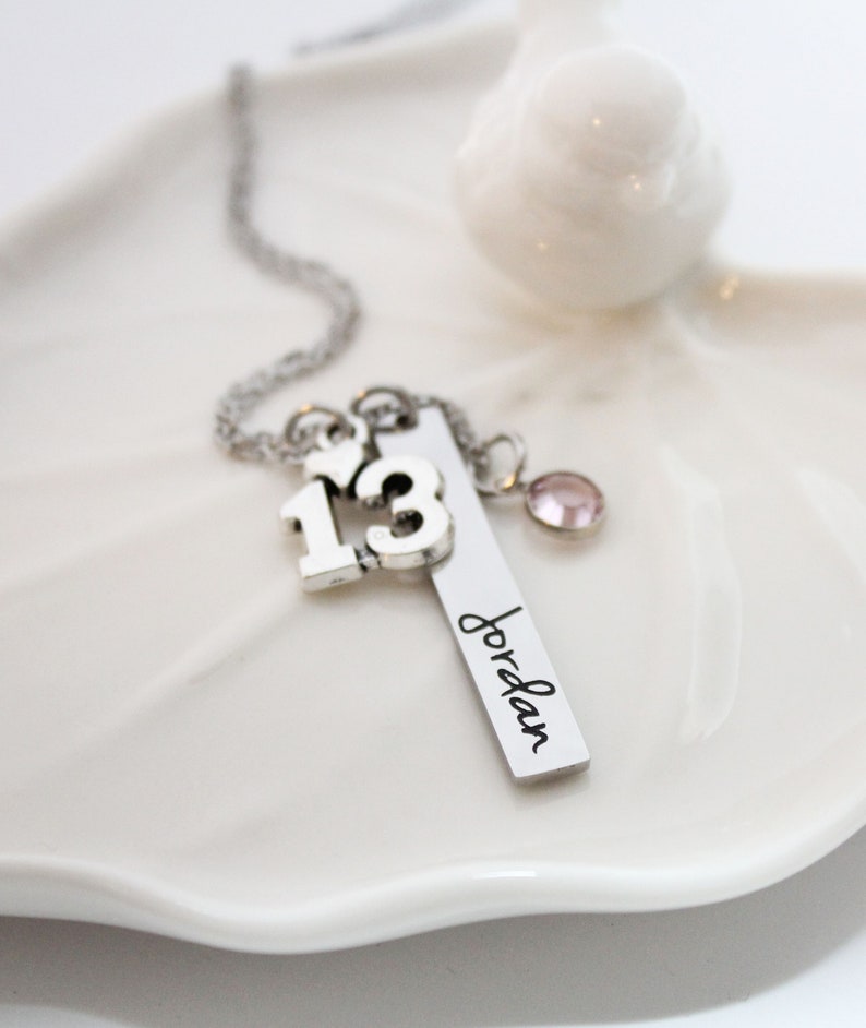 Puede incluir: Collar de plata con un dije que dice "13" y un dije con el nombre "Jordan" grabado. El collar tambi&eacute;n tiene un peque&ntilde;o dije de cristal rosa.