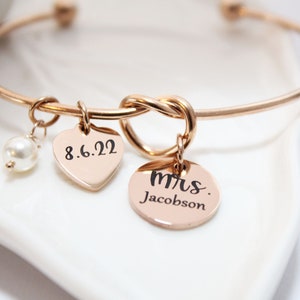 May include: A rose gold bangle bracelet with a knotted design and three charms. The charms are a heart with the date 8.6.22, a pearl, and a round charm with the text "Mrs. Jacobson".