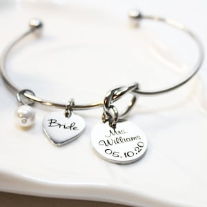May include: A silver bangle bracelet with a knot charm, a heart charm with the word "Bride" engraved on it, a pearl charm, and a round charm with the text "Mrs. Williams 05.10.20" engraved on it.
