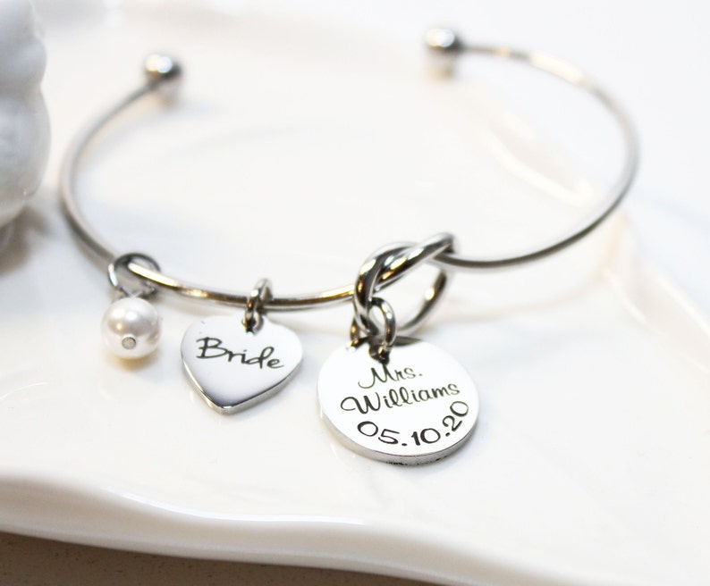 May include: A silver bangle bracelet with a knot charm, a heart charm with the word "Bride" engraved on it, a pearl charm, and a round charm with the text "Mrs. Williams 05.10.20" engraved on it.
