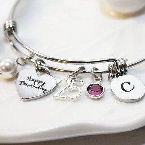 May include: Silver charm bracelet with a heart charm that says "Happy Birthday", a charm with the number 25, a small purple gemstone charm, and a round charm with the letter C.