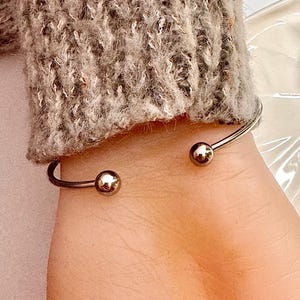 Puede incluir: Una pulsera de pu&ntilde;o plateada con una peque&ntilde;a bola dorada en cada extremo abierto. La pulsera se lleva en una mu&ntilde;eca, con una manga de punto gris visible en el fondo.