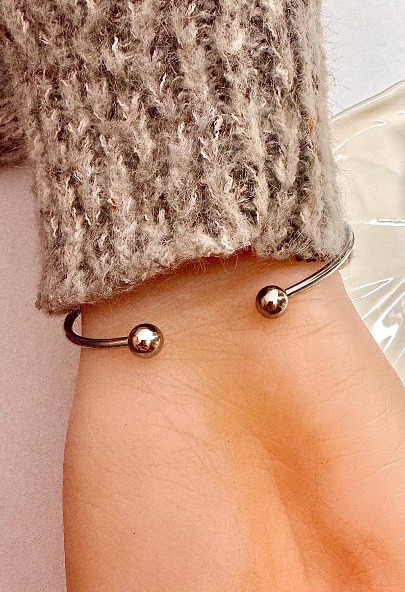 Puede incluir: Una pulsera de pu&ntilde;o plateada con una peque&ntilde;a bola dorada en cada extremo abierto. La pulsera se lleva en una mu&ntilde;eca, con una manga de punto gris visible en el fondo.