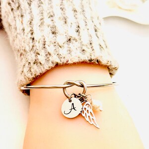 Puede incluir: Pulsera de brazalete plateada con dise&ntilde;o de nudo. Cuenta con un dije redondo con la letra "A", un dije de ala y una peque&ntilde;a perla. La pulsera se muestra en una mu&ntilde;eca, con un su&eacute;ter de color neutro en el fondo.