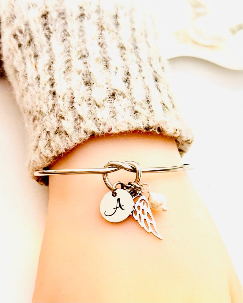 Puede incluir: Pulsera de brazalete plateada con dise&ntilde;o de nudo. Cuenta con un dije redondo con la letra "A", un dije de ala y una peque&ntilde;a perla. La pulsera se muestra en una mu&ntilde;eca, con un su&eacute;ter de color neutro en el fondo.