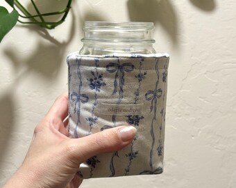 May include: A clear glass jar with a blue and white floral patterned cozy. The cozy features a ribbon and floral design. The text "odette and co" is printed on the cozy. Green leaves are in the background.