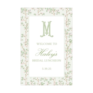 May include: A rectangular sign with a floral border and a central panel. The sign reads "Welcome to Haley's Bridal Luncheon" with the date 5.30.25. The border features a light green background with pink flowers and green branches.