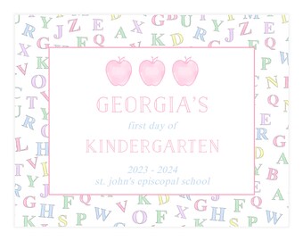 May include: A pink and white first day of kindergarten sign with a pastel alphabet border. The sign says "Georgia's First Day of Kindergarten 2023-2024 St. John's Episcopal School". There are three pink apples with green stems and leaves at the top of the sign.